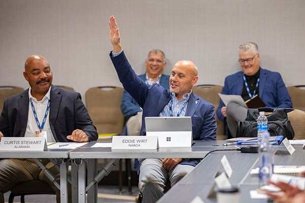 Four men smiling with two sitting in the front behind a table as one has his hand raised. The other two are in the background.