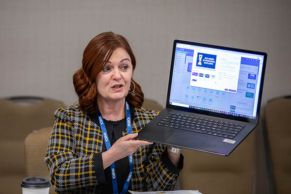 Woman with red hair, dressed in a checkered jacket and a blue lanyard holding a laptop and talking.
