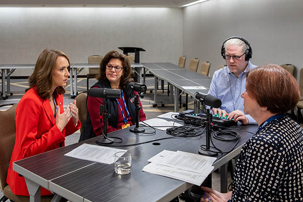 Three women and a man sit at a small table with audio recording equipment, while one woman in a red suit jacket speaks into a microphone.