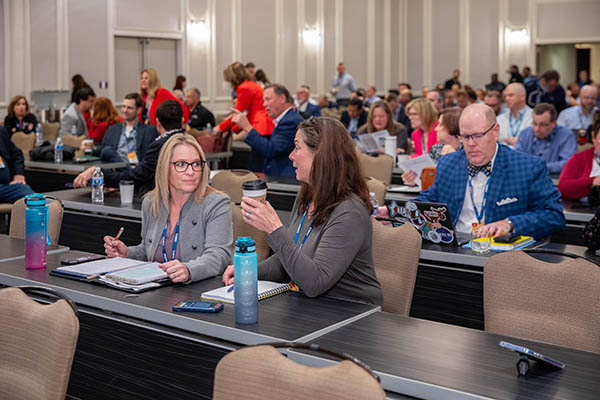 Conference room filled with people sitting at tables and talking. Focus on two women having a personal, private conversation. 