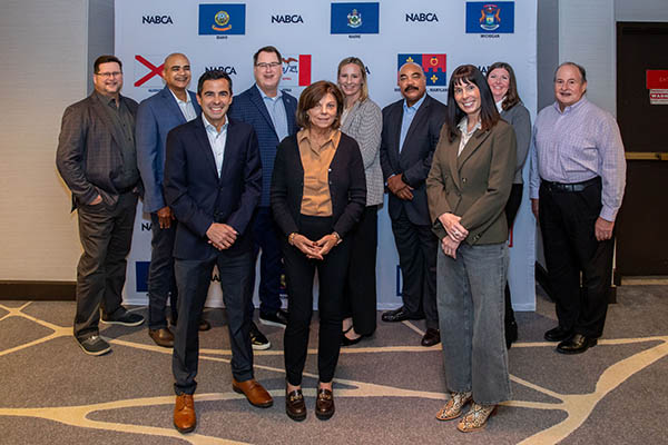 Six men and four women in business attire standing and smiling in front of a trade show banner depicting 16 US state flags and one county flag.