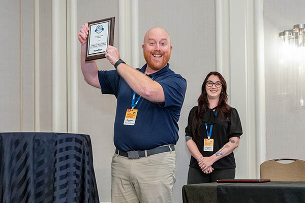 A man in a blue shirt and khakis holds an award on stage, smiling, while a woman in a dark shirt and glasses smiles in the background.